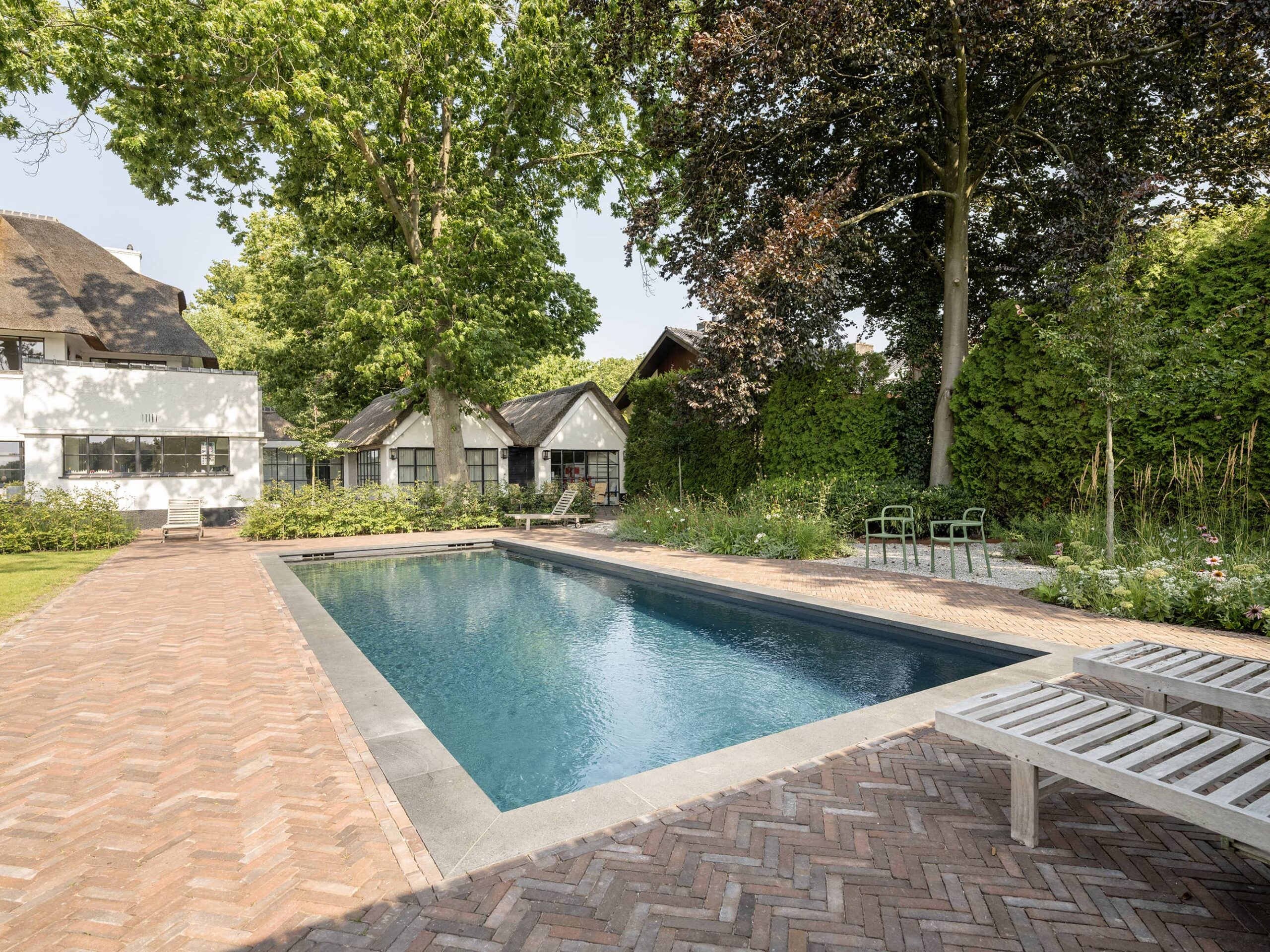 moderne zwembad in ruime groene tuin met visgraat bestrating-compressed Modern buitenzwembad in ruime groene tuin met visgraat bestrating, ligbedden, volwassen bomen en witte villa op de achtergrond.